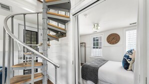 Une chambre avec un escalier en colimaçon en bois, un lit recouvert d’un couvre-lit à motifs et une horloge murale ronde.