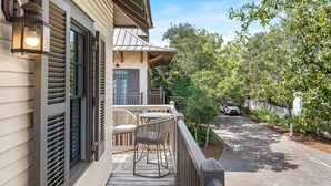 Une terrasse en bois, dotée de mobilier d’extérieur, d’une rambarde et offrant une vue sur la rue, avec des arbres et une voiture garée.