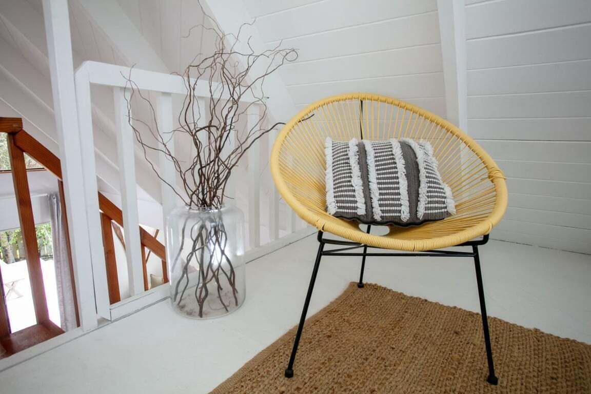Una sedia in vimini con un cuscino a righe, un vaso di vetro con dei rami e una ringhiera in legno.
