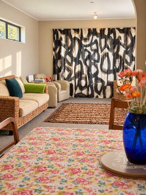 A living room with a sofa, a coffee table with a floral tablecloth, a wicker chair, and a decorative vase with flowers. - Simply Sophia (Rotorua)