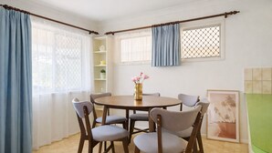 Una zona de menjador amb una taula rodona i sis cadires, una finestra amb cortines i una estant amb objectes decoratius.