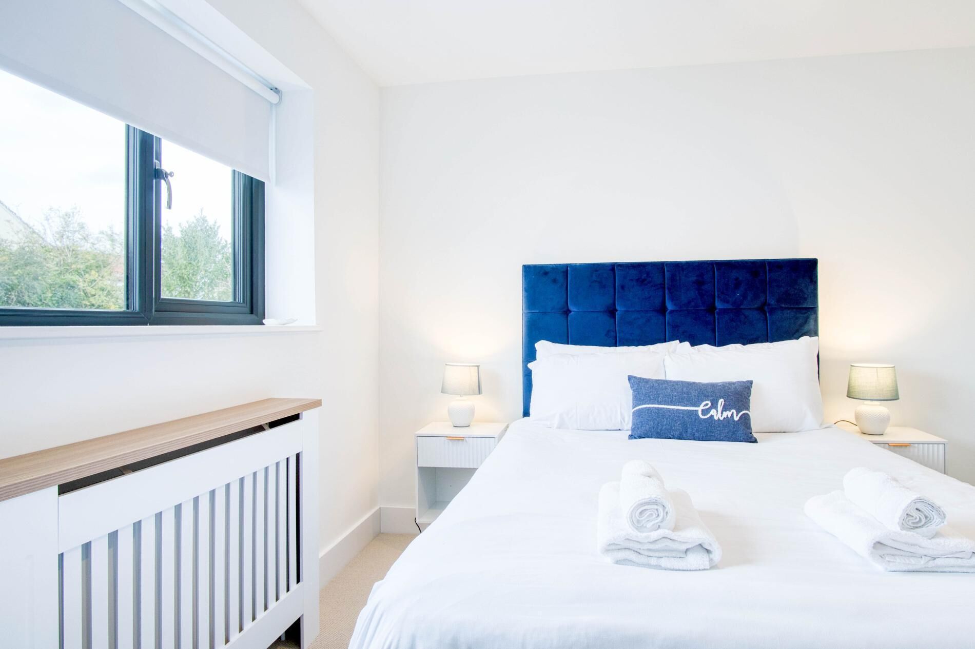 A neatly made bed with white linens and two rolled towels, a blue headboard, and two bedside lamps.