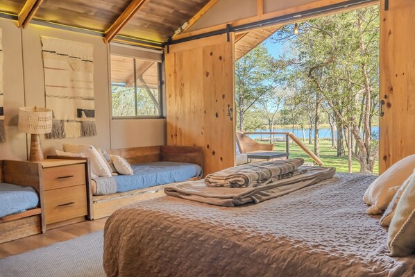 A cozy bedroom with a large bed, two smaller beds, a wooden nightstand, a lamp, and a view of a lake and trees. - GLAT Austin (Bastrop)