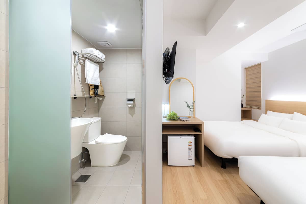 Ein modernes Hotelzimmer mit Toilette, Waschbecken und einem kleinen Kühlschrank. Es gibt ein Bett mit weißer Bettwäsche und einem Holzkopfstück. Ein Spiegel hängt an der Wand, und auf dem Nachttisch steht eine Pflanze.
