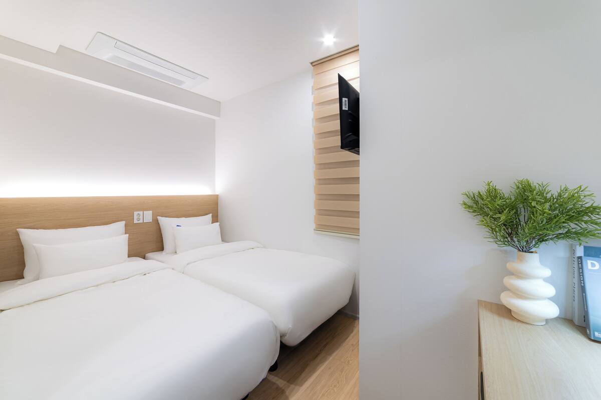 Ein Hotelzimmer mit zwei Betten, einem an einer Holzplatte befestigten Flachbildfernseher, einer kleinen Topfpflanze und einem Nachttisch mit einem Buch.