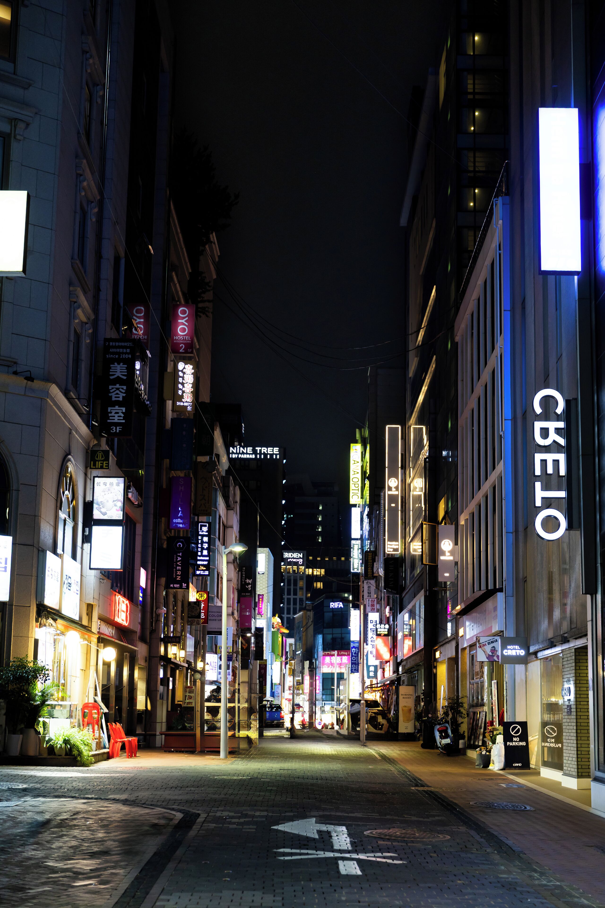 Vista nocturna de una calle con letreros y escaparates iluminados, incluyendo un hotel llamado "CBELO" a la derecha.