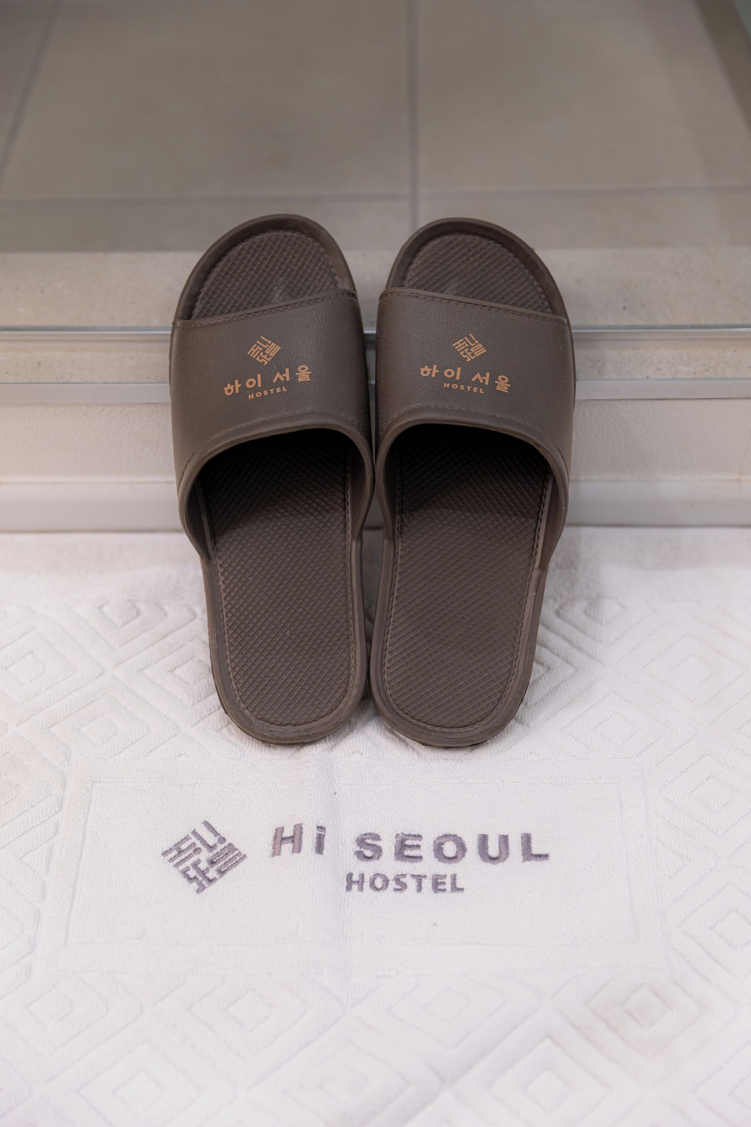 Braune Hausschuhe mit dem Schriftzug "Hi Seoul Hostel" auf der Sohle, platziert auf einem gemusterten Boden.