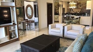 A modern living room with a large black ottoman, white sofas, a glass partition, and a dining area with a chandelier. - Garza Blanca Resort & Spa Los Cabos (Cabo San Lucas)