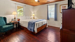Una camera da letto con soffitto in legno, un letto con coperta blu, un comodino con lampada, una poltrona verde e un armadio in legno.