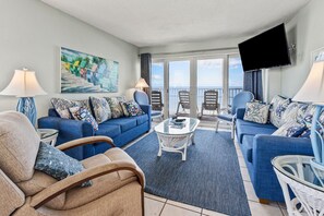 A spacious living room with a large window, a flat-screen TV, and a balcony with chairs. - Oceanfront 3BR condo in Cherry Grove with pool and private balcony views (North Myrtle Beach)