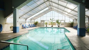 Une piscine intérieure à l’eau d’un bleu limpide, entourée de chaises bleues et de plantes en pot, le tout sous une structure de toit en verre.