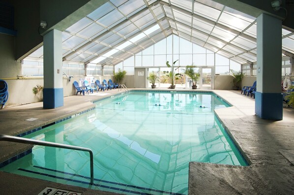An indoor swimming pool with clear blue water, surrounded by blue chairs and potted plants, under a glass roof structure. - Oceanfront 3BR condo in Cherry Grove with pool and private balcony views (North Myrtle Beach)