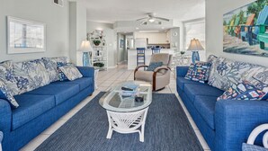 Un salon avec deux canapés bleus, une table basse en verre et un fauteuil en osier blanc. Au fond, on aperçoit une cuisine avec des meubles blancs et un coin repas.