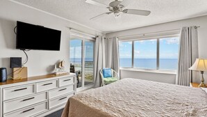 Une chambre avec un grand lit, un téléviseur fixé au mur, un ventilateur de plafond et une vue sur la mer par les fenêtres.