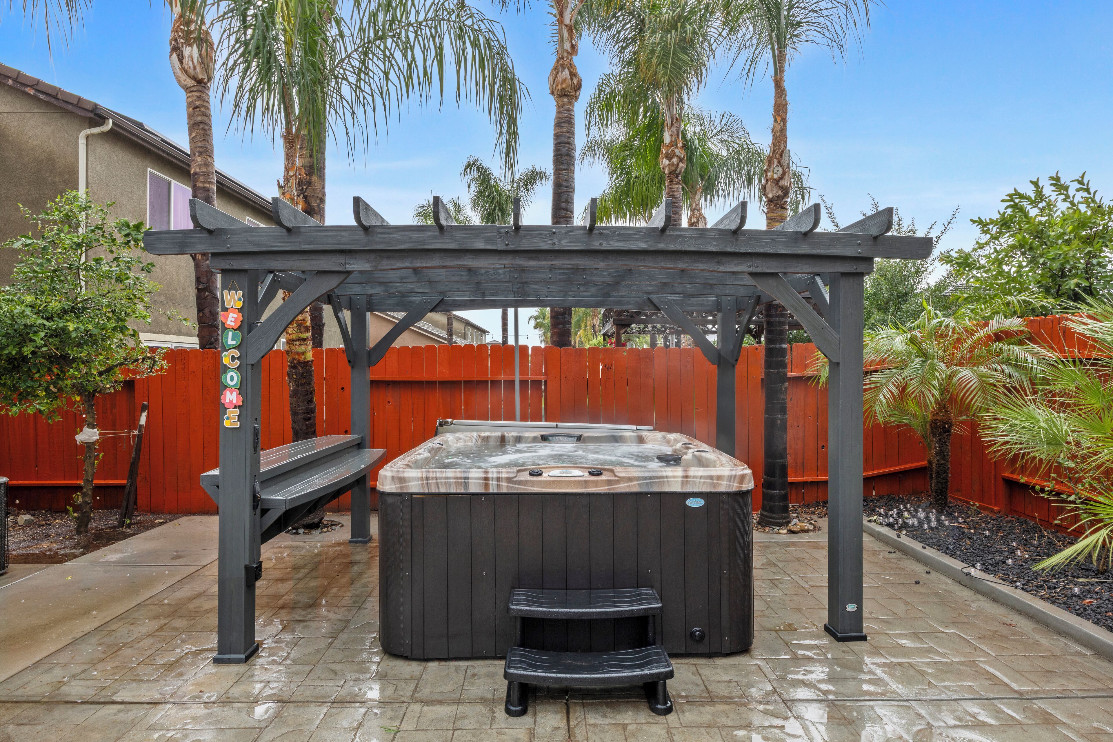 A hot tub under a wooden pergola with a wooden bench in front, surrounded by palm trees and a red fence.