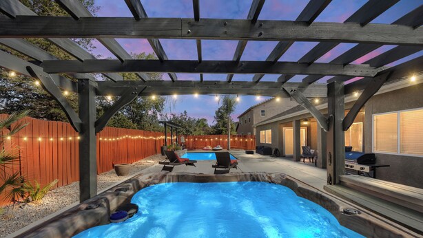 A hot tub under a wooden pergola with string lights, surrounded by a wooden fence and a barbecue area. - Luxury Escape on Mondego Dr – Pool, Spa & Pool Table (Bakersfield)