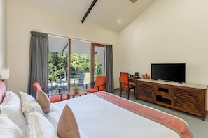 A hotel room with a large bed, a TV on a wooden cabinet, a balcony with chairs, and a view of greenery.