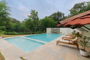 Una piscina rectangular con agua azul cristalina, rodeada por una terraza de piedra. Hay sillas de descanso de madera con toallas, y un pequeño edificio con techo rojo al fondo.