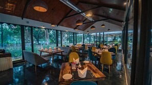 A restaurant with tables and chairs, large windows, and a view of trees. - Aranya Vilas - A Nature Resort (Udaipur)