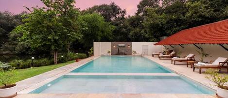 A rectangular swimming pool with a stone border, surrounded by a grassy area and trees. There are lounge chairs and a covered area with a red roof.