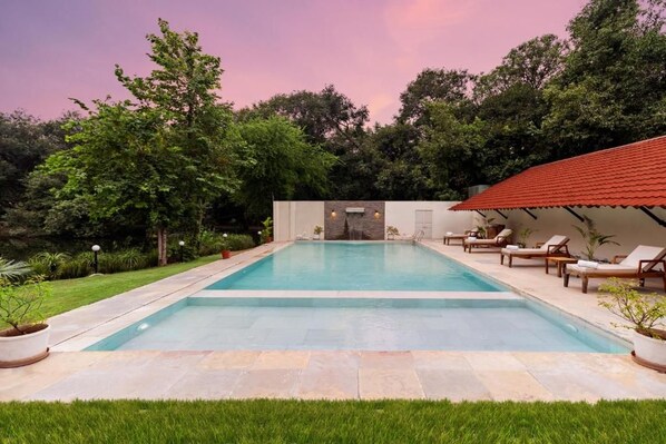 A rectangular swimming pool with a stone border, surrounded by a grassy area and trees. There are lounge chairs and a covered area with a red roof. - Aranya Vilas - A Nature Resort (Udaipur)