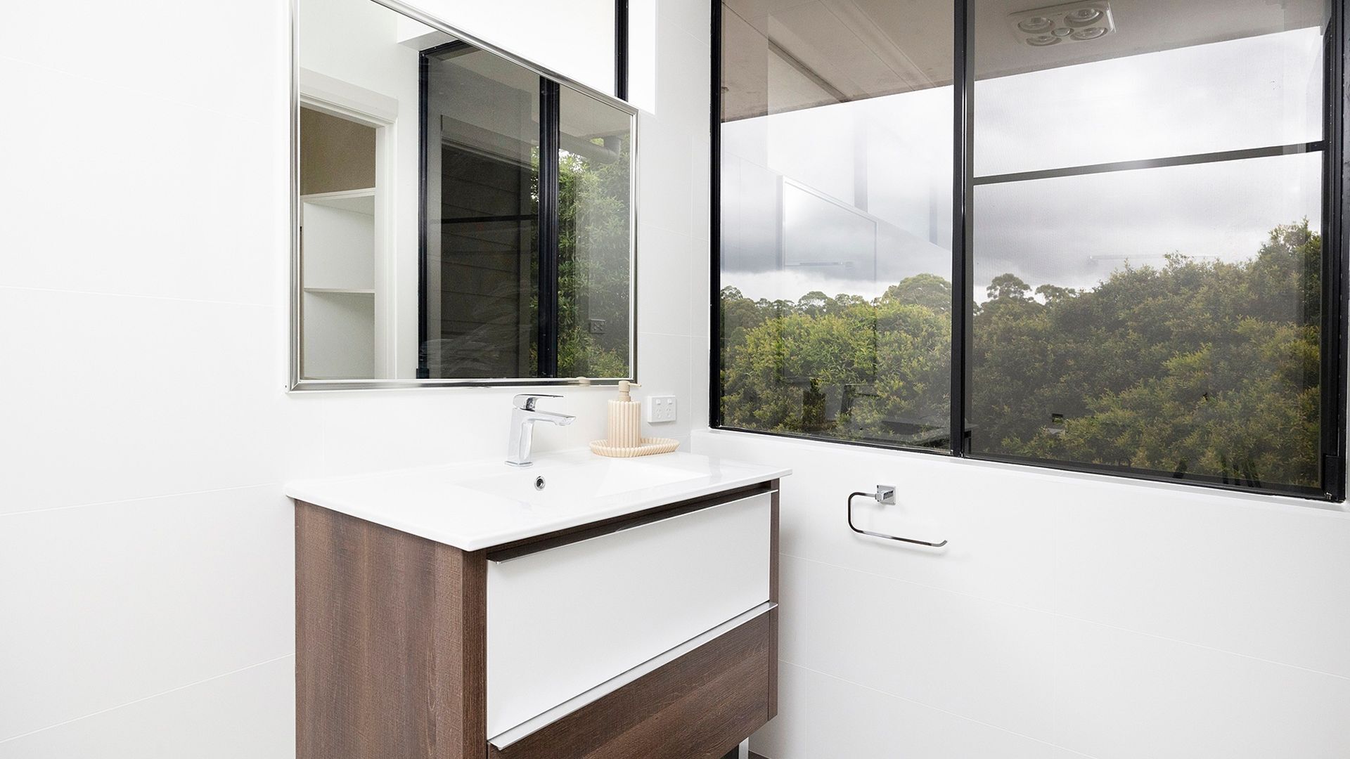 A modern bathroom with a white vanity, a large mirror, and a window offering a view of a forested area.