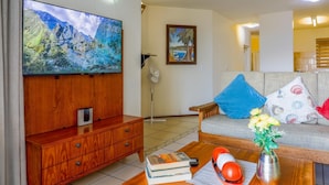 Ein Wohnzimmer mit einem Holzkabinett, einem Flachbildfernseher, einer Couch mit gemusterten Kissen, einem Couchtisch mit Büchern und Dekorationsgegenständen sowie einem Gemälde an der Wand.