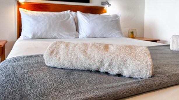Ein ordentlich gemachtes Bett mit grauer Decke, gerolltem Handtuch, weißen Kissen und einem gerahmten Bild am Kopfteil.
