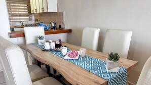 Zona pranzo con tavolo in legno, tovaglia fantasia, sedie bianche e un piccolo vaso con pianta.