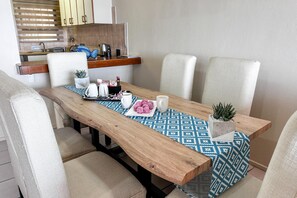 Un comedor con mesa de madera, mantel estampado, sillas blancas y una pequeña planta en maceta.