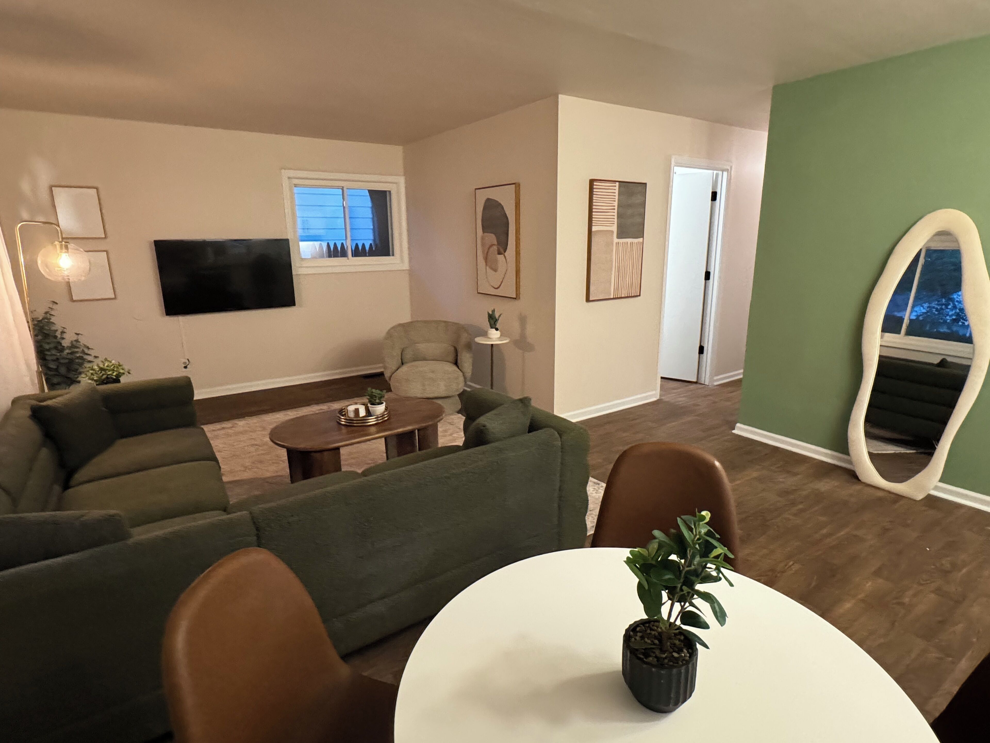 A living room with a green sofa, a coffee table, and a potted plant. There is a flat-screen TV mounted on the wall and a door leading to another room.