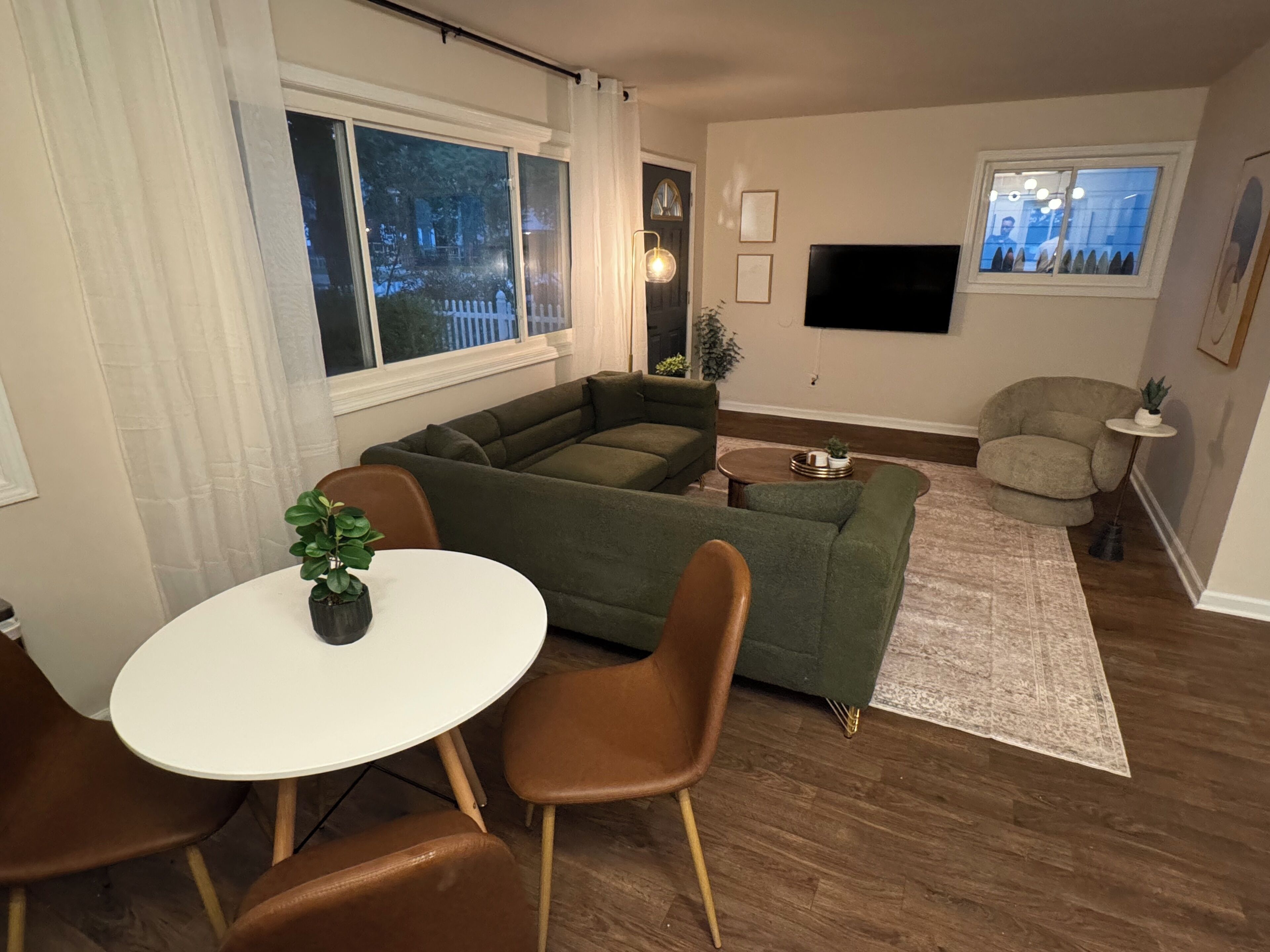 A living room with a sectional sofa, a round dining table with chairs, a small side table, a TV mounted on the wall, and a window with curtains.