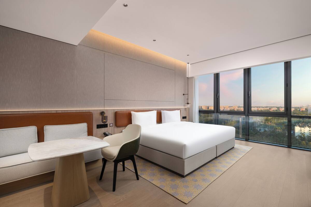 Habitació d'hotel moderna amb un llit gran, un sofà, una cadira i una taula petita. L'habitació té un finestral gran amb vista a la ciutat.