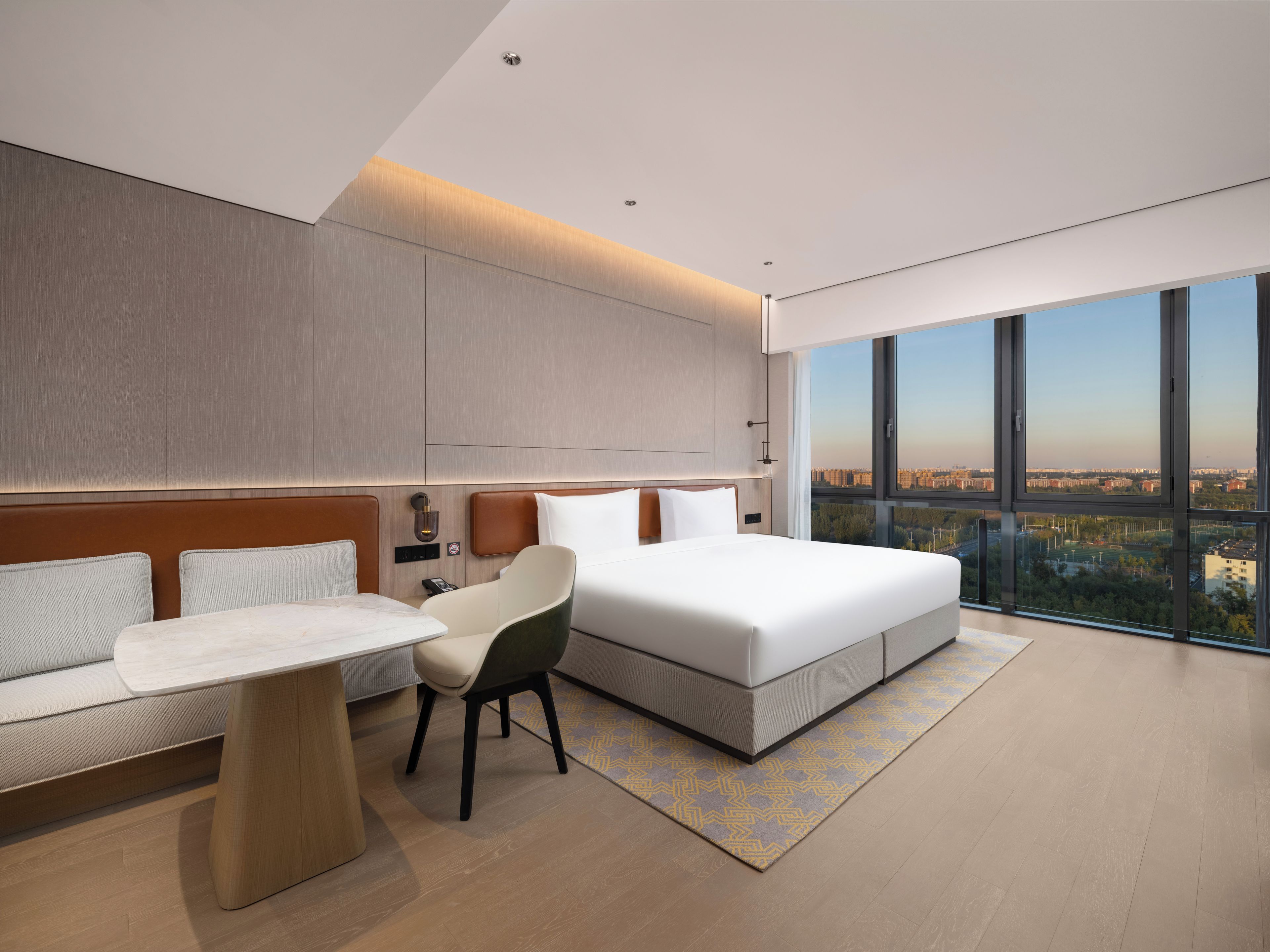 Une chambre d’hôtel moderne avec un grand lit, un canapé, un fauteuil et une petite table. Une grande fenêtre offre une vue sur la ville.