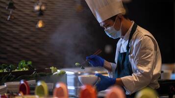 A chef in a white chef's hat and apron, wearing a face mask, preparing food in a kitchen with a modern design and a brick wall background.