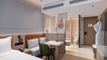 A modern hotel room with a bed, bedside table, green chair, desk, and a bathroom visible through an open door.