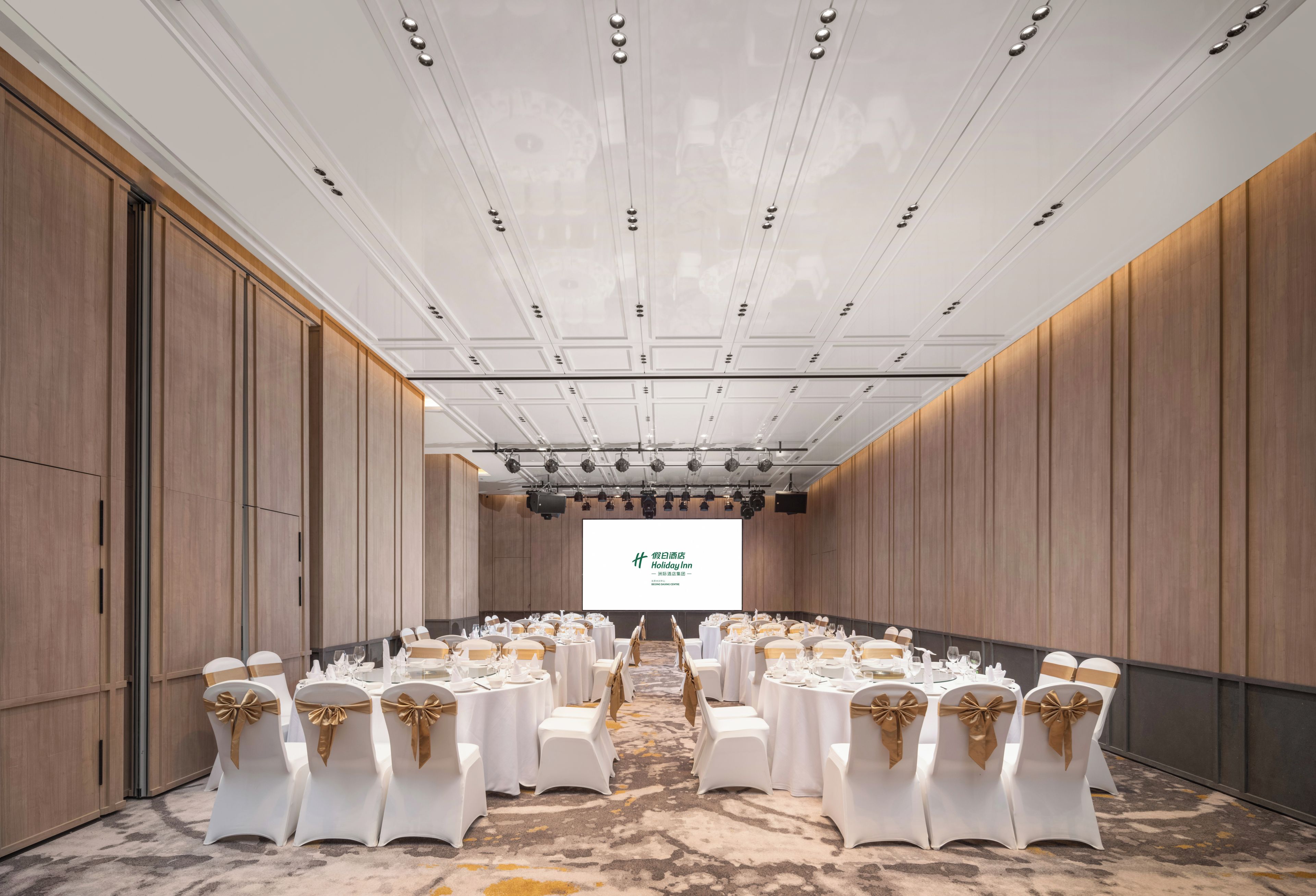 A conference room with white tables and chairs, a large screen at the front, and wooden paneling on the walls.