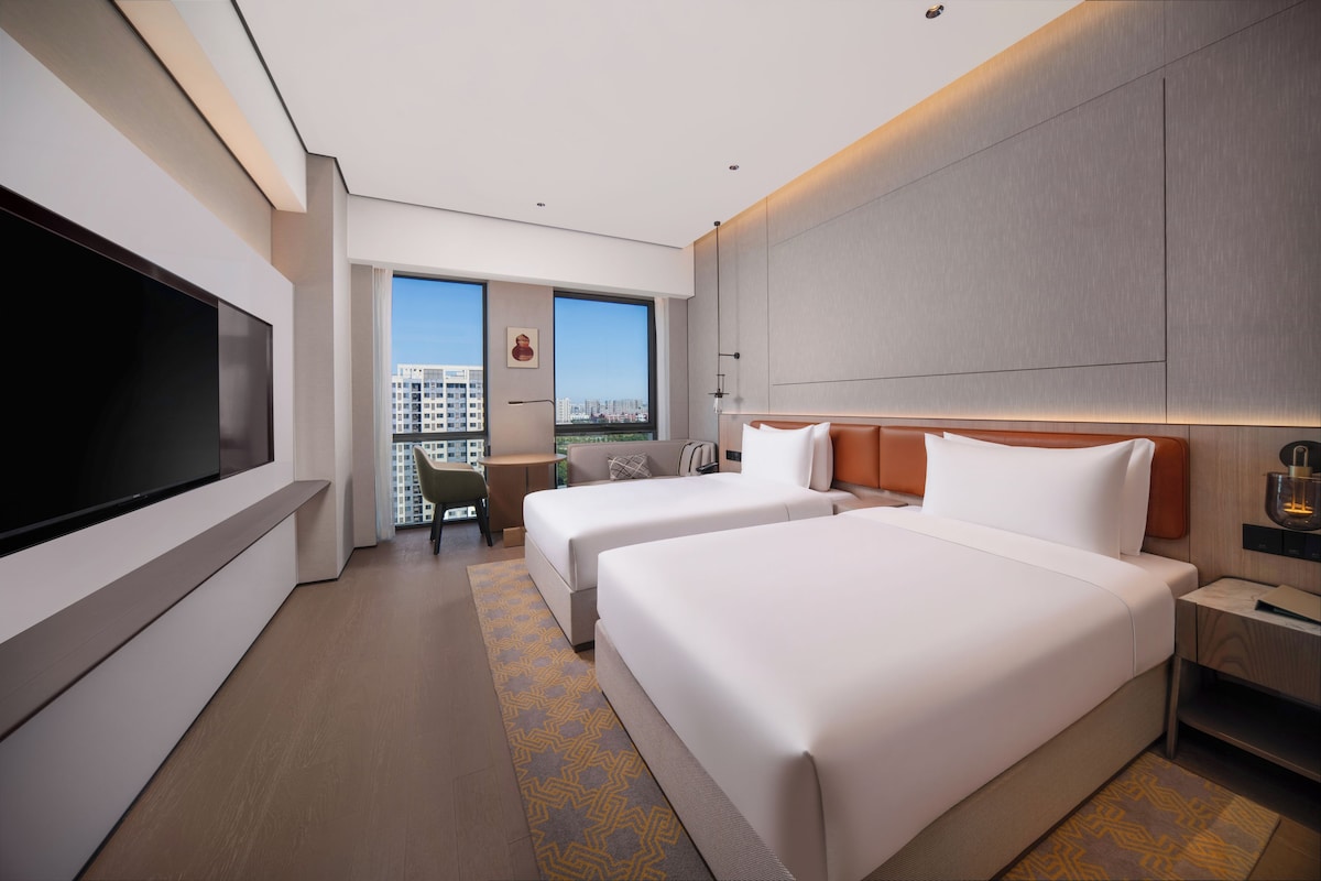 A hotel room with two beds, a TV, a desk, and a view of the city.
