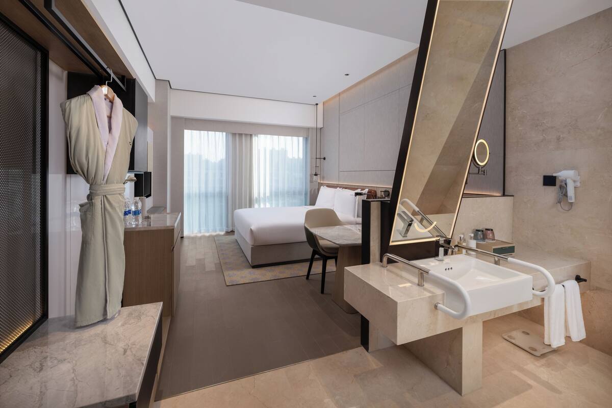 Una habitació d'hotel moderna amb un llit gran, una zona de seients, un bany amb un mirall gran i un lavabo, i una bata penjada en un suport.