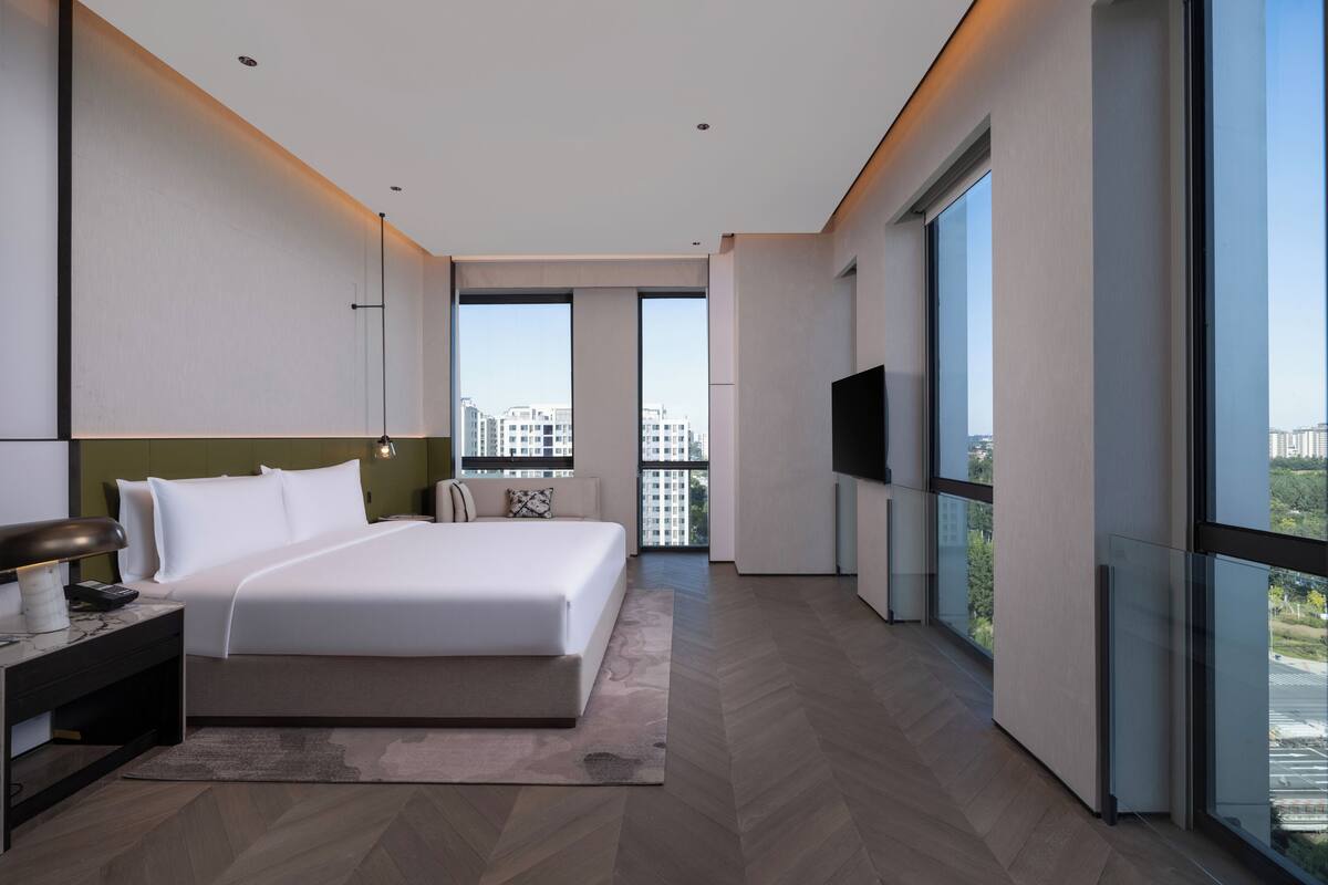Habitació d'hotel moderna amb un llit gran, una taula de nit, una televisió i vistes a la ciutat a través de grans finestres.