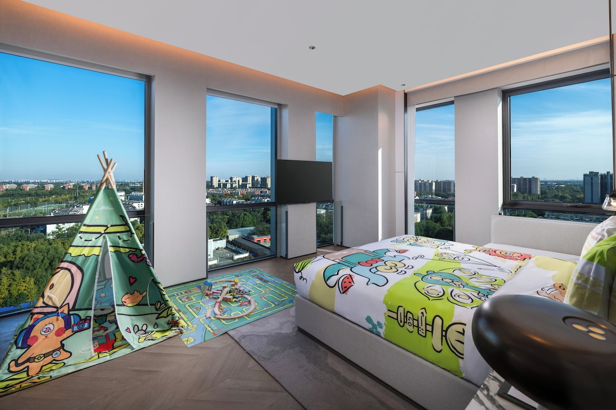 A modern bedroom with large windows, a bed with cartoon-themed bedding, a play mat, and a teepee.