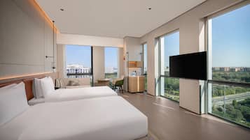 A modern hotel room with two large beds, a desk, a chair, a TV, and large windows offering city views.