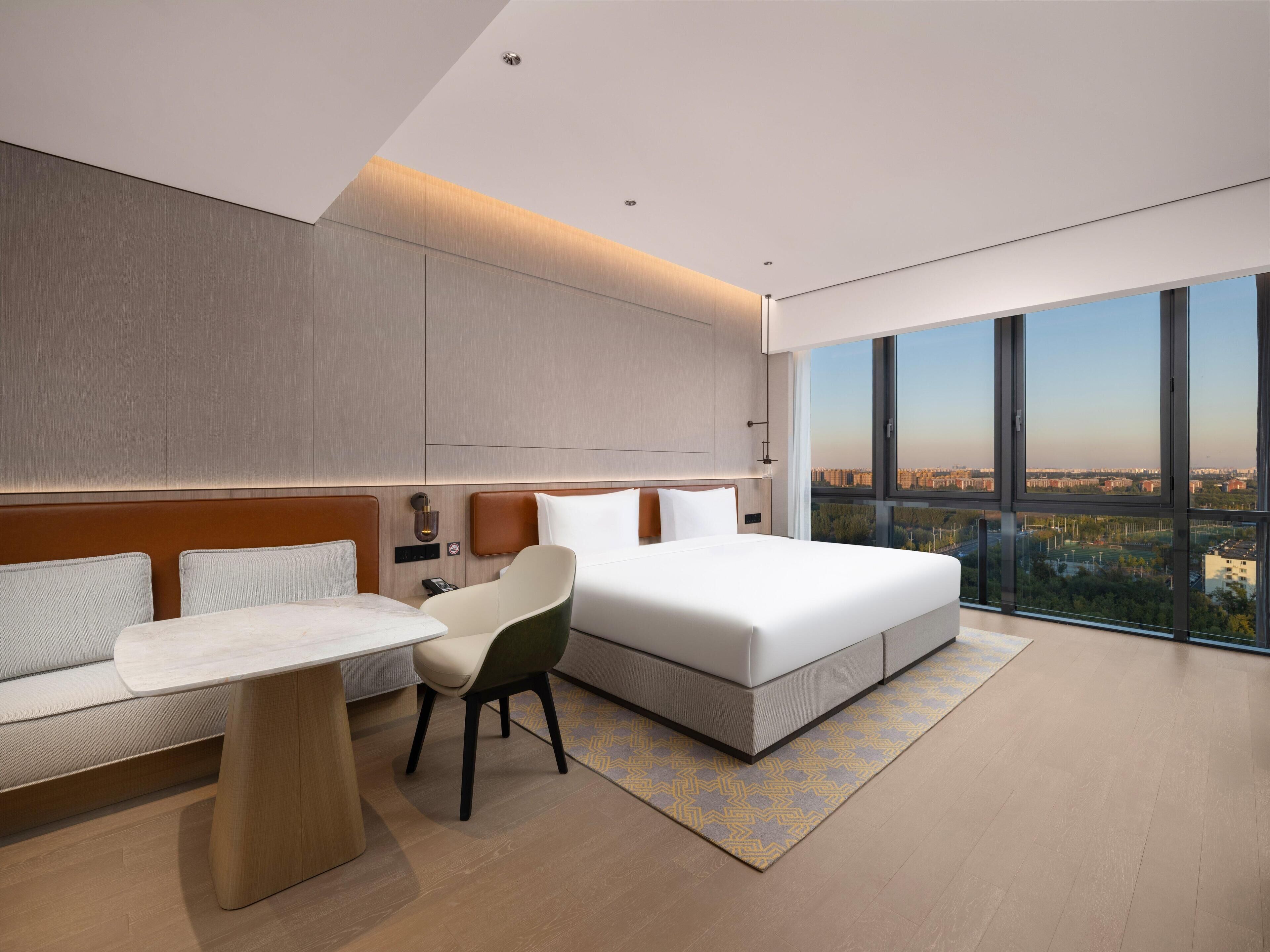 Una habitación de hotel moderna con una cama grande, un sofá, una silla y un escritorio. Una ventana amplia ofrece vistas a la ciudad.