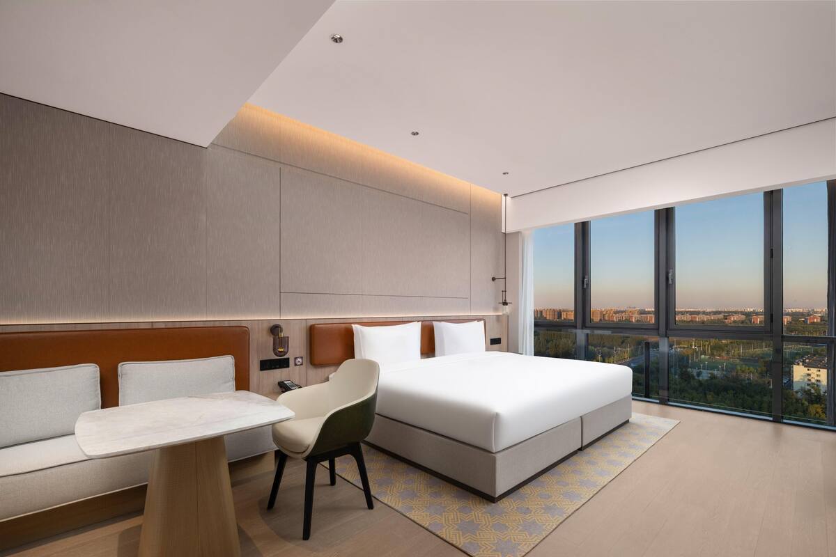 Habitació d'hotel moderna amb un llit gran, un sofà, una cadira i un escriptori. Hi ha un gran finestral que ofereix vistes a la ciutat.