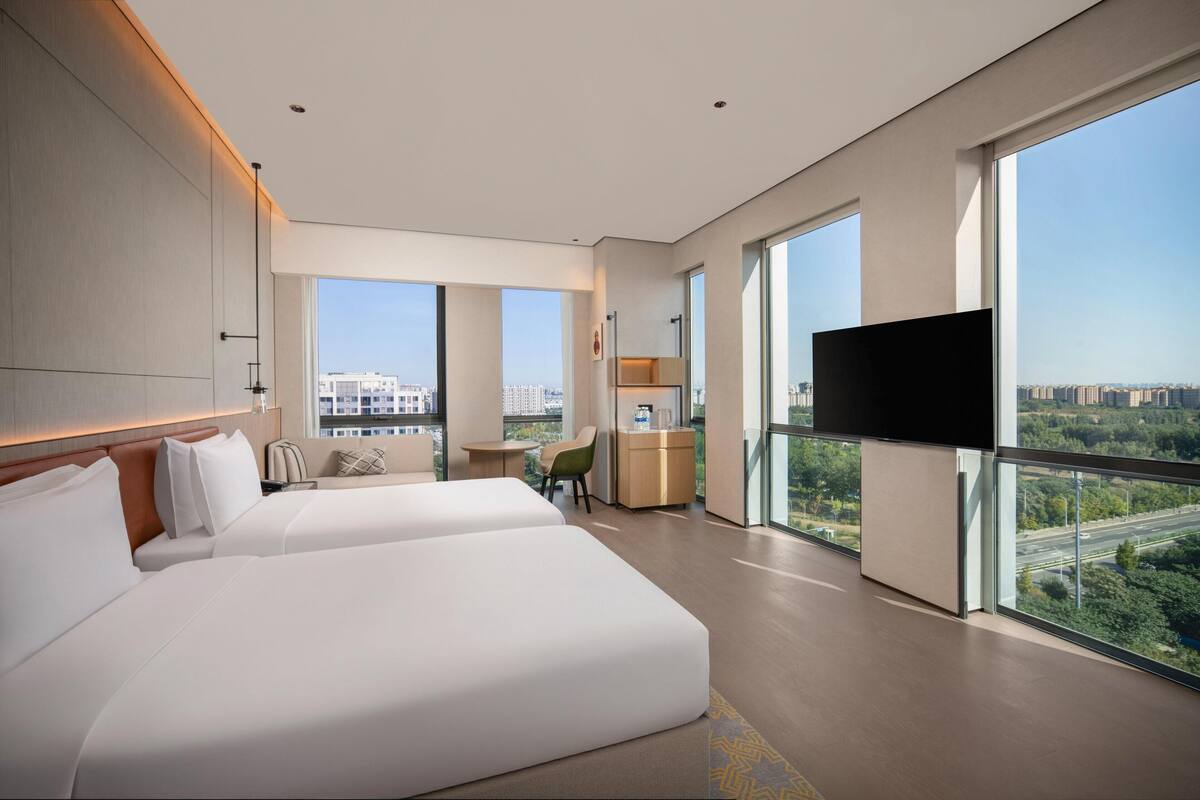 Habitació d'hotel moderna amb dos llits grans, escriptori, cadira, televisió i grans finestres que donen vistes a la ciutat.