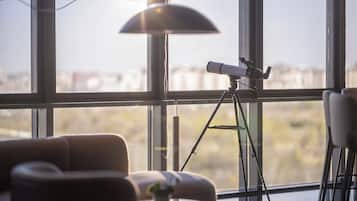 A room with a telescope on a tripod, large windows, a lamp, and a chair.