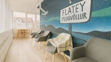 Uma área de espera com um mural, cadeiras cinzas, uma mesa e uma placa que diz "FLATEY FLUGVÖLLUR".