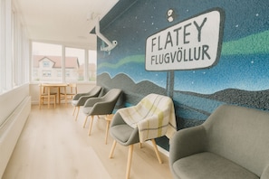 Un área de espera con un mural, sillas grises, una mesa y un letrero que dice "FLATEY FLUGVÖLLUR".