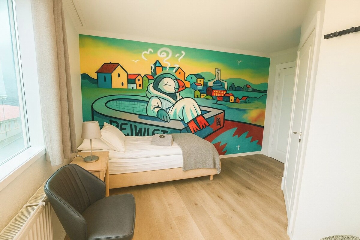 Ein Schlafzimmer mit einem Bett, einem Sessel, einem Nachttisch, einer Lampe und einem Wandbild.