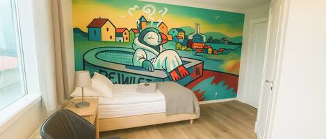 A bedroom with a bed, a chair, a nightstand, a lamp, and a wall mural.